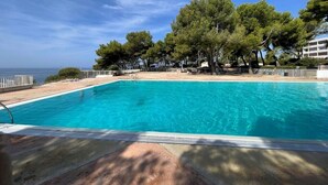 A clear blue pool with a wooden deck in the foreground, surrounded by trees and a view of the sea.