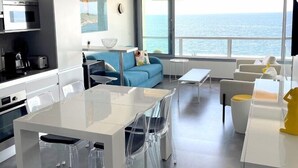 A modern living room with a large window overlooking the sea, a dining area with a white table and chairs, and a kitchen with modern appliances.
