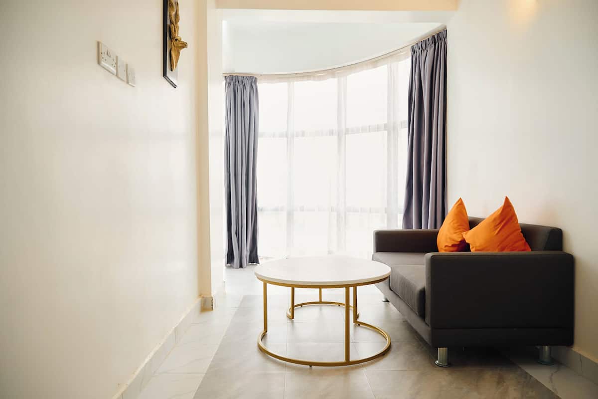 A modern living room with a round table, a sofa with orange cushions, and a large window with curtains.