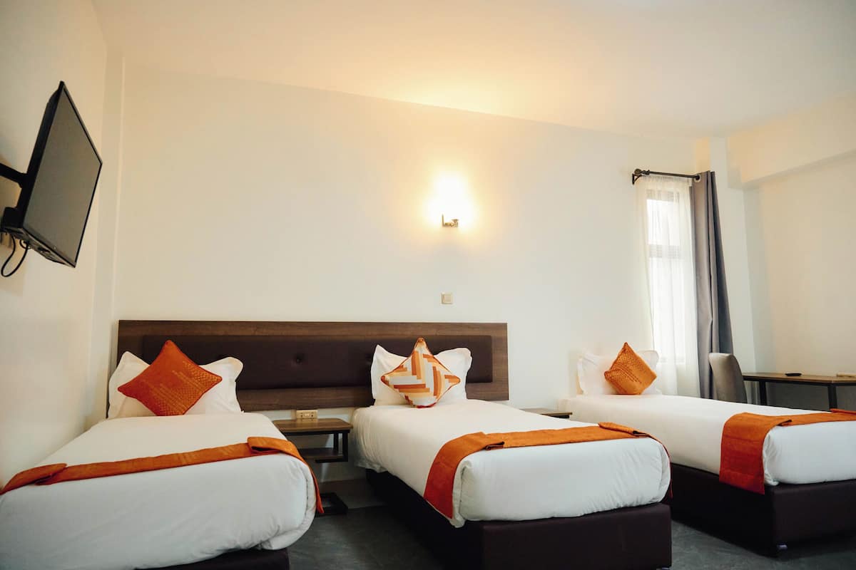 A hotel room with three beds, a TV mounted on the wall, a desk, and a window with curtains.