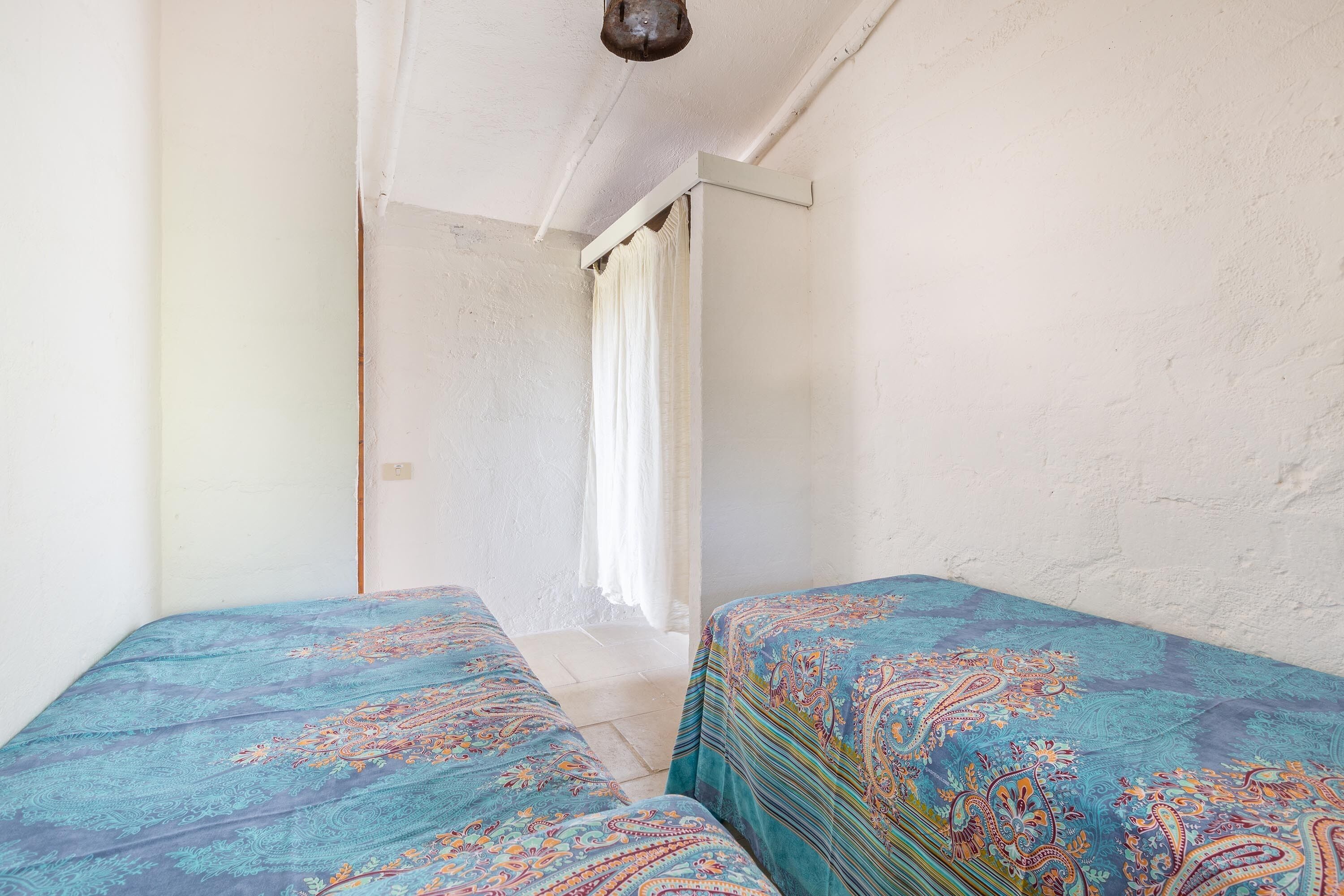 Una stanza con due letti, pareti bianche, una finestra con tende bianche e una lampada a soffitto.