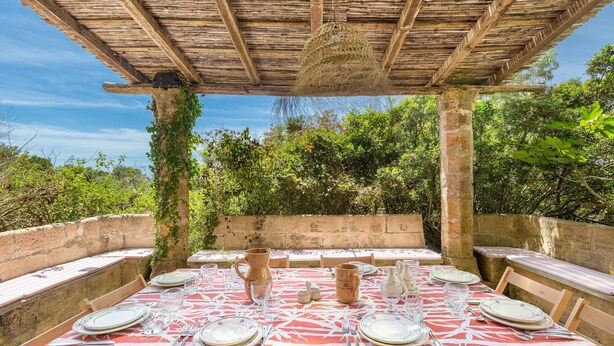 Un espace extérieur aménagé pour les repas, avec une pergola en bois rustique, des murs en pierre et une végétation luxuriante en arrière-plan.