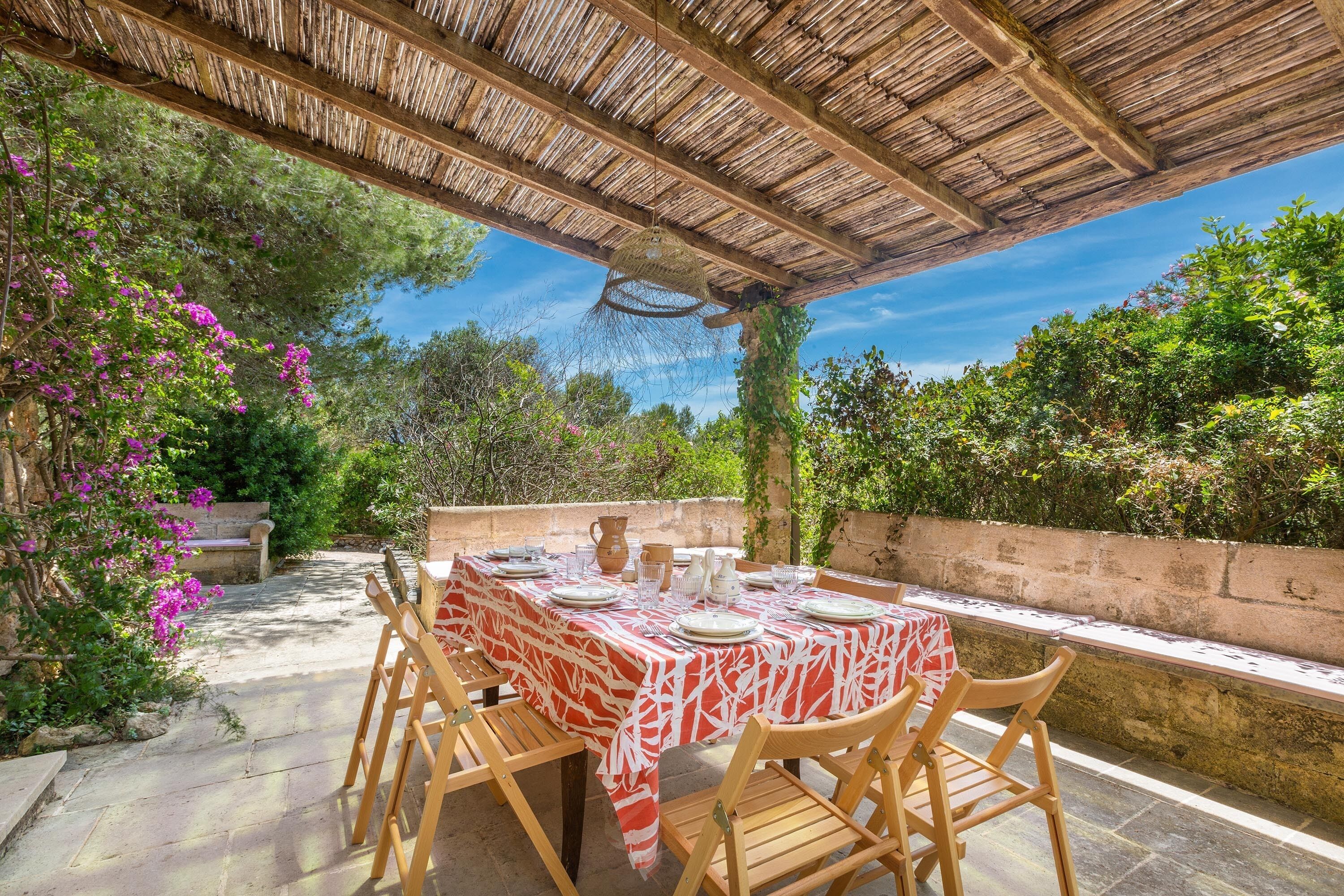 An outdoor dining area with a table set for four, surrounded by wooden chairs, under a thatched roof.