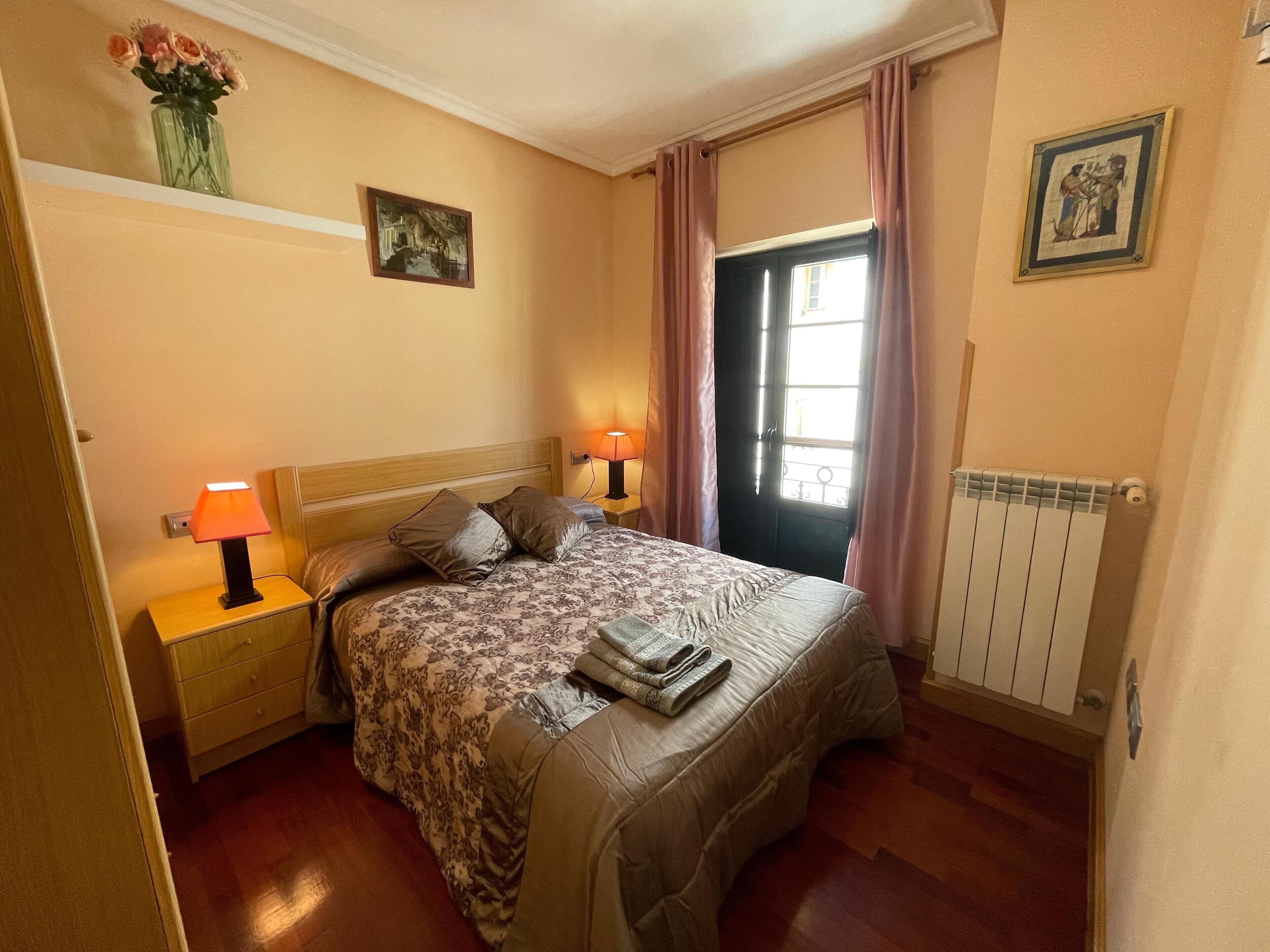 Ein Schlafzimmer mit einem Bett, zwei Nachttischlampen, einer Heizung, einem Fenster mit Vorhängen und einem Wandbild.
