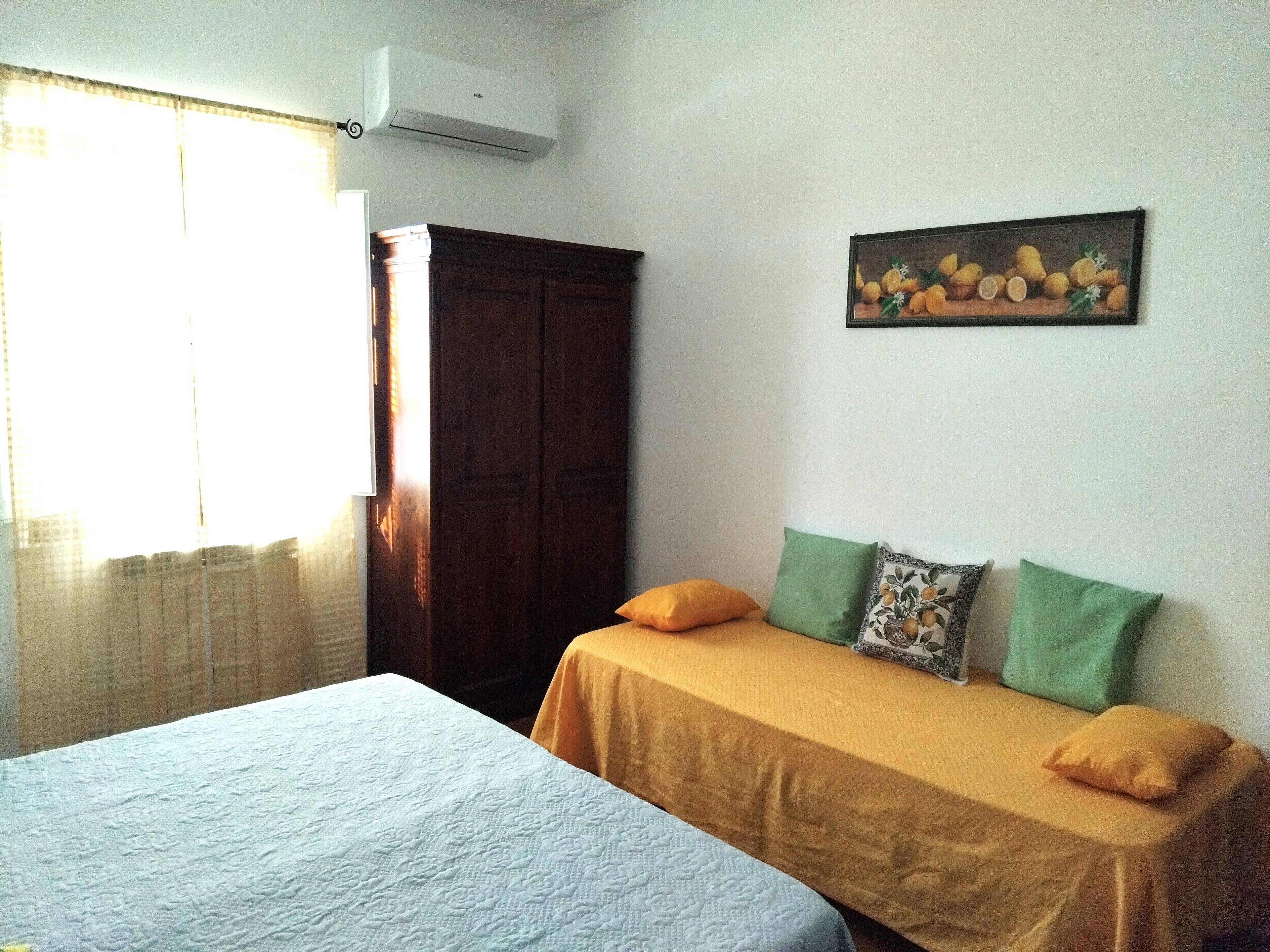 A room with two beds, a wooden wardrobe, a painting of fruit, and an air conditioning unit.