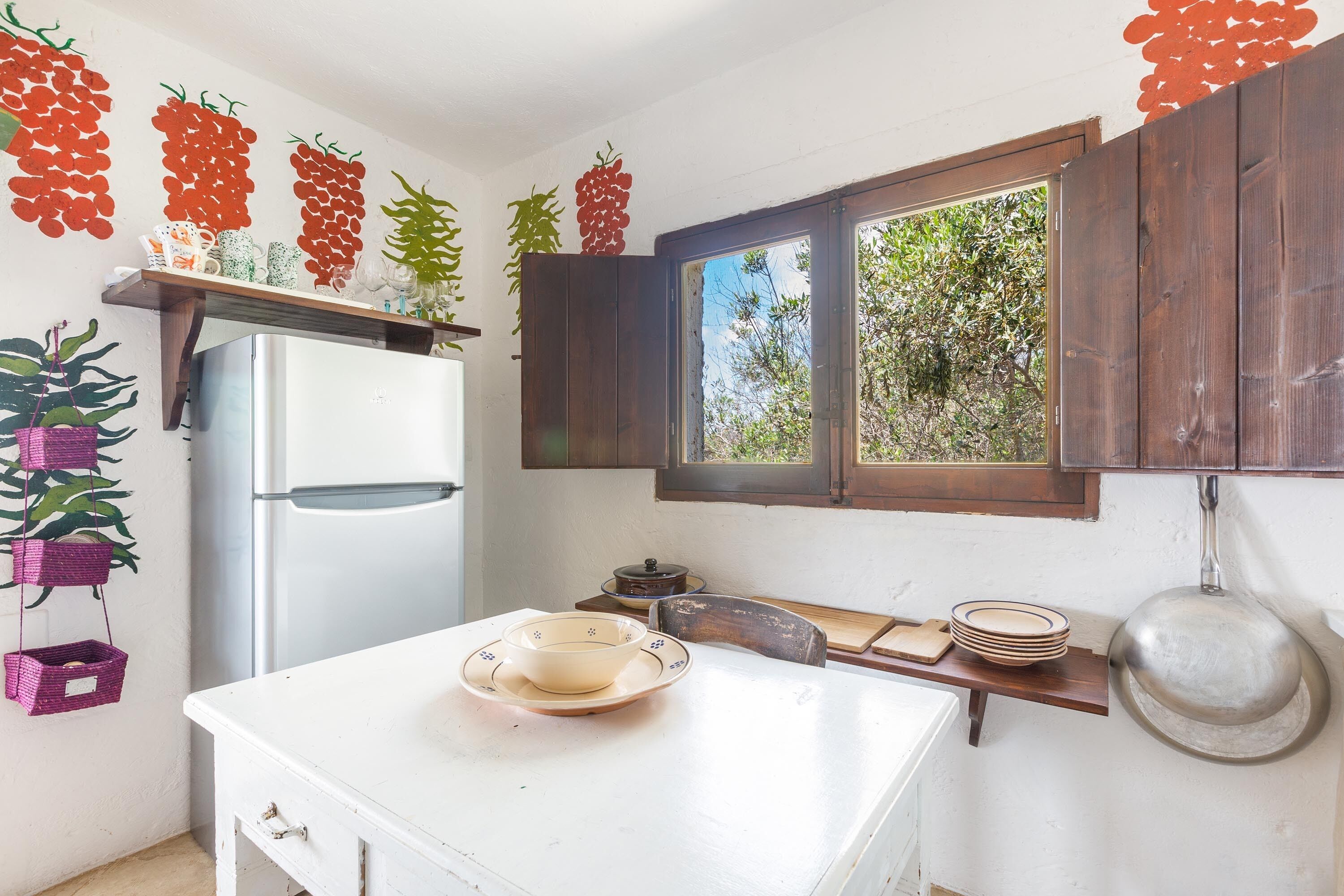 A kitchen with a white refrigerator, wooden cabinets, a window with a view of greenery, and a shelf with decorative items.