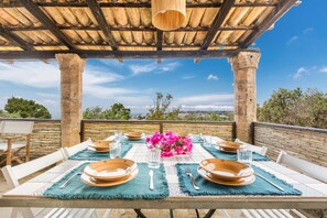 An outdoor dining area with a table set for four, under a wooden pergola with a clear view of the landscape.