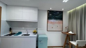 A compact kitchen with white cabinets, a small fridge, and a sink. There's a painting on the wall and a chair near a window with curtains. - Vista mar no Costa Azul (Costa Azul Salvador)
