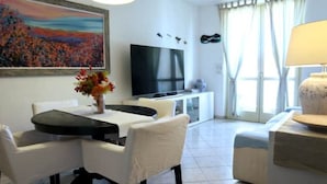 Een moderne woonkamer met een eethoek, een flatscreen televisie en een groot raam met gordijnen.