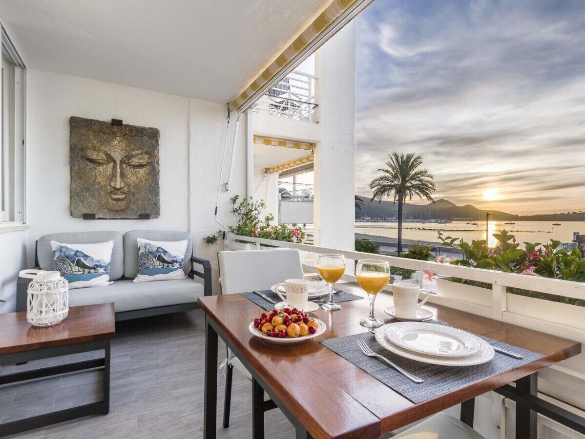 A balcony with a dining table set for breakfast, a sofa with cushions, and a view of the sea at sunset.