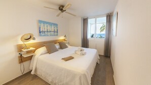 Une chambre d’hôtel avec un lit, un ventilateur de plafond, un tableau représentant des voiliers, une fenêtre avec des rideaux et une vue sur la mer.