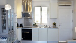 Une cuisine moderne avec des meubles blancs, une hotte de cuisinière en acier inoxydable, un four à micro-ondes et un lave-vaisselle. On y trouve une fenêtre avec des stores, une porte et un climatiseur mural.