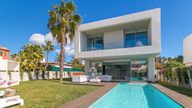Et moderne hus med to etager, en swimmingpool, hvide havemøbler og palmer.