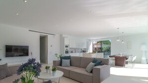 En moderne stue med sofa, sofabord og TV. Der er et køkkenområde med skabe og et spisebord.