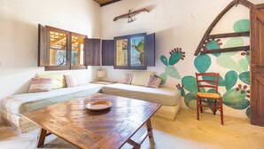 A cozy room with a wooden table, a cushioned sofa, and a chair. There are windows with wooden frames, a wall with a cactus mural, and a wooden door. - 3504 PJE - Casa Fico D'india by Barbarhouse (Castrignano del Capo)