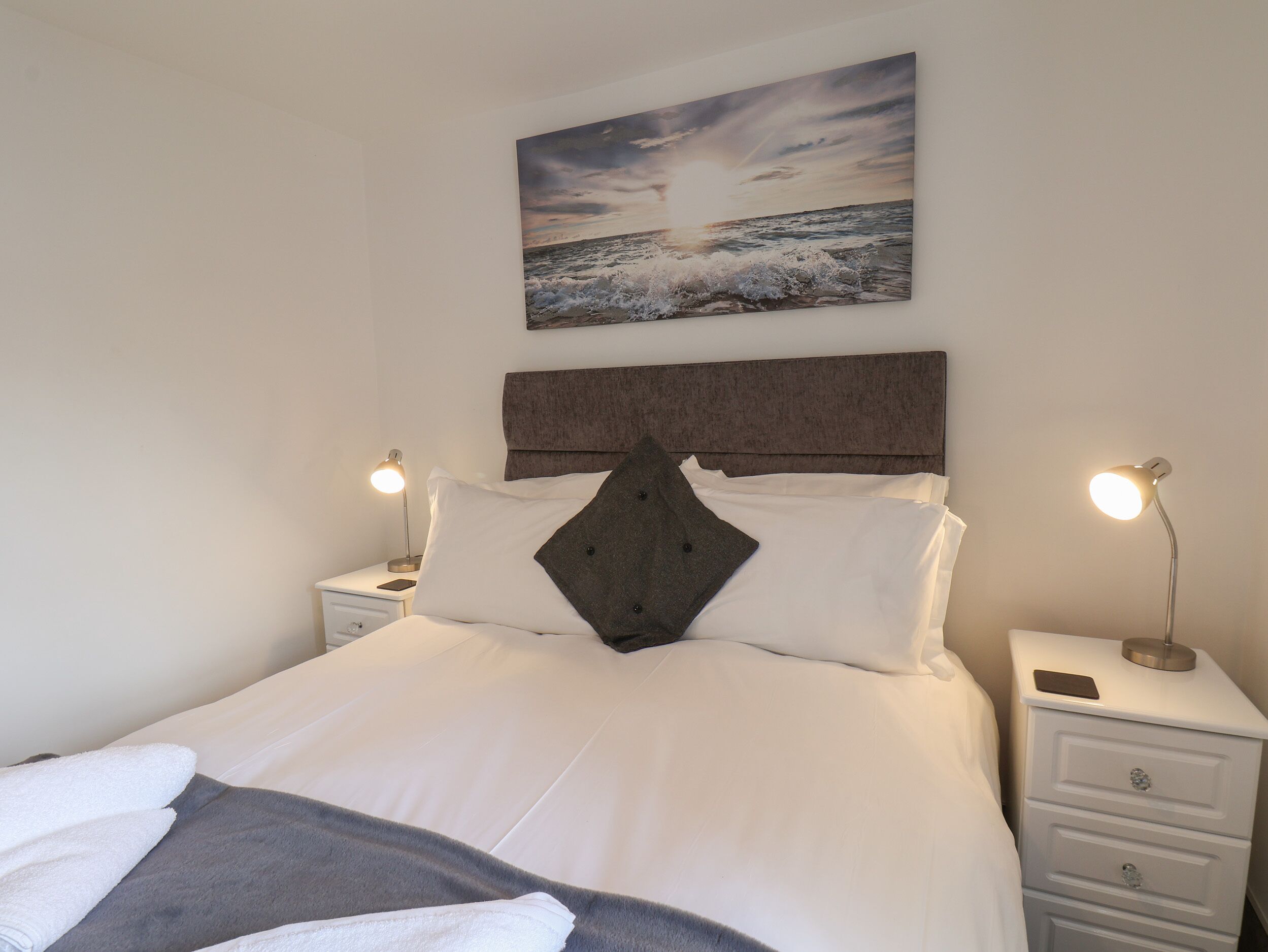 A neatly made bed with white linens and a gray throw blanket, flanked by two bedside tables with lamps and a smartphone on each. A painting of a sunset over the ocean hangs above the bed.