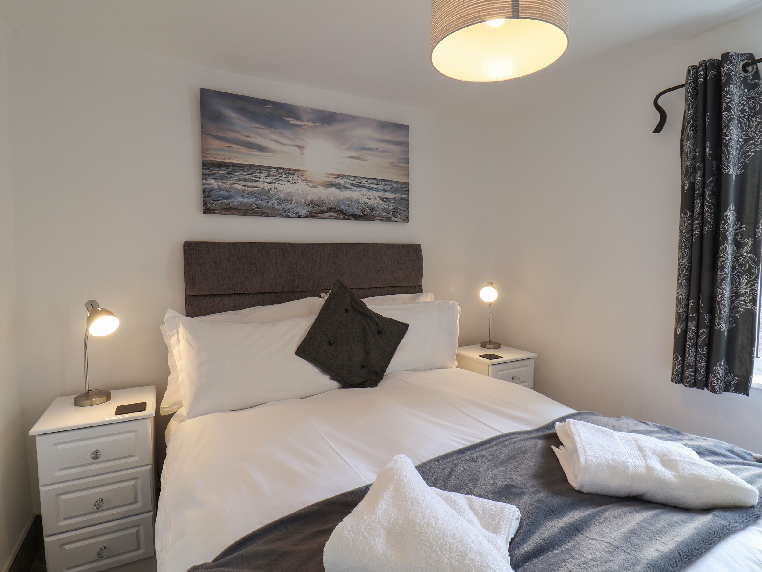 A neatly arranged bedroom with a bed, bedside lamps, a nightstand, a wall-mounted painting, and a window with curtains.