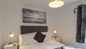 A neatly arranged bedroom with a bed, bedside lamps, a nightstand, a wall-mounted painting, and a window with curtains.