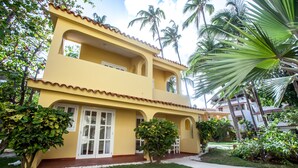 A two-story yellow building with a balcony, surrounded by greenery and palm trees. - BEACH VIEW PREMIUM 1BR SUITE Ground Floor - Los Corales Beach Village #25 (Punta Cana)