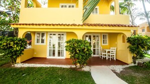 A two-story yellow building with a patio area, white doors, and a small seating area with chairs. - BEACH VIEW PREMIUM 1BR SUITE Ground Floor - Los Corales Beach Village #25 (Punta Cana)