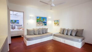 A modern room with a white sofa set, a bed with a view, and two framed beach paintings. - BEACH VIEW PREMIUM 1BR SUITE Ground Floor - Los Corales Beach Village #25 (Punta Cana)