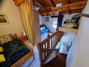 A bedroom with a bed, a wooden headboard, a window with curtains, a small table with a lamp, and a view of a blue sofa in the adjacent room. - Maisonnette du Clos - Avec piscine partagée (Argeles-sur-Mer)