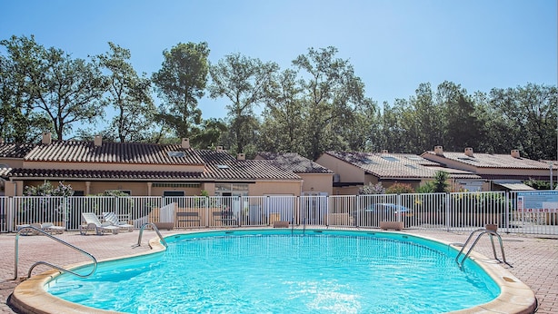 A swimming pool with a tiled deck, surrounded by a white fence, and adjacent to a row of buildings with similar architecture.