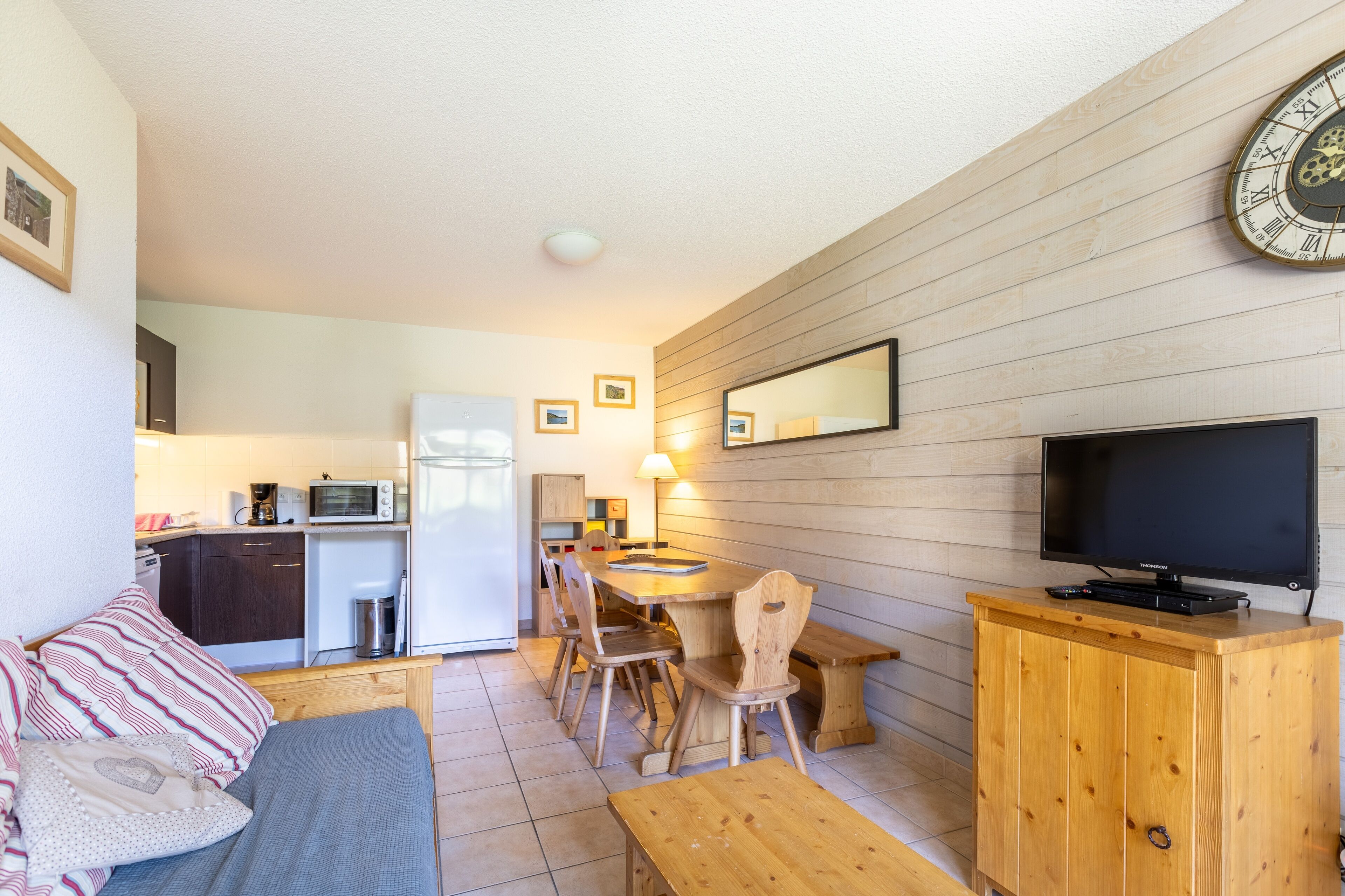 A compact living space with a kitchenette, a dining area with wooden furniture, a television on a wooden cabinet, and a wall-mounted clock.