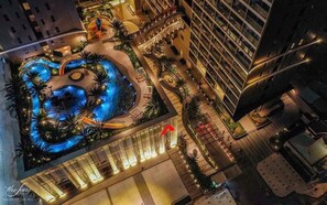 Vue aérienne d’un hôtel doté d’une grande piscine illuminée, entourée de palmiers et bordée d’une passerelle incurvée.