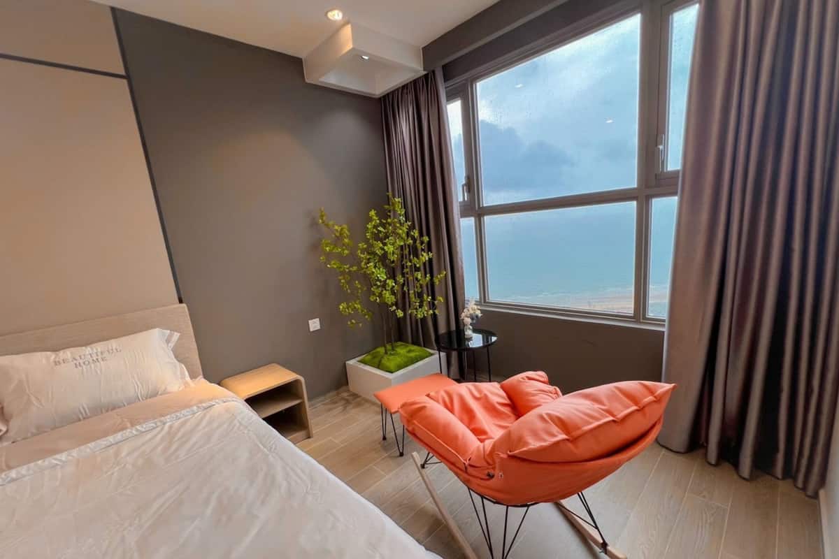 Une chambre d’hôtel moderne, équipée d’un lit, d’une petite table avec une plante, d’une chaise et d’une grande fenêtre avec des rideaux.