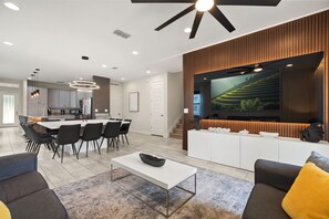 A modern living room with a flat-screen TV, a dining area with a white table and chairs, and a kitchen in the background.