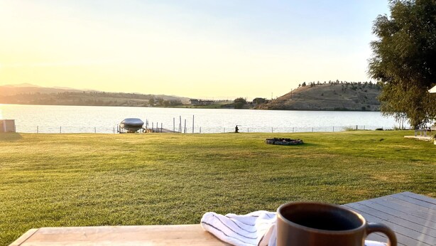Une table en bois, avec une tasse et une serviette, donnant sur un lac, avec une zone herbeuse et une colline en arrière-plan.