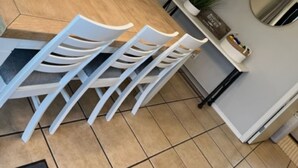 Un coin repas avec une table ronde en bois entourée de chaises blanches, des cartes encadrées accrochées au mur et une plante en pot posée sur une étagère.