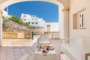 Una terraza con mobiliario de ratán blanco, una mesa con alimentos para el desayuno y una vista de un edificio blanco al fondo.