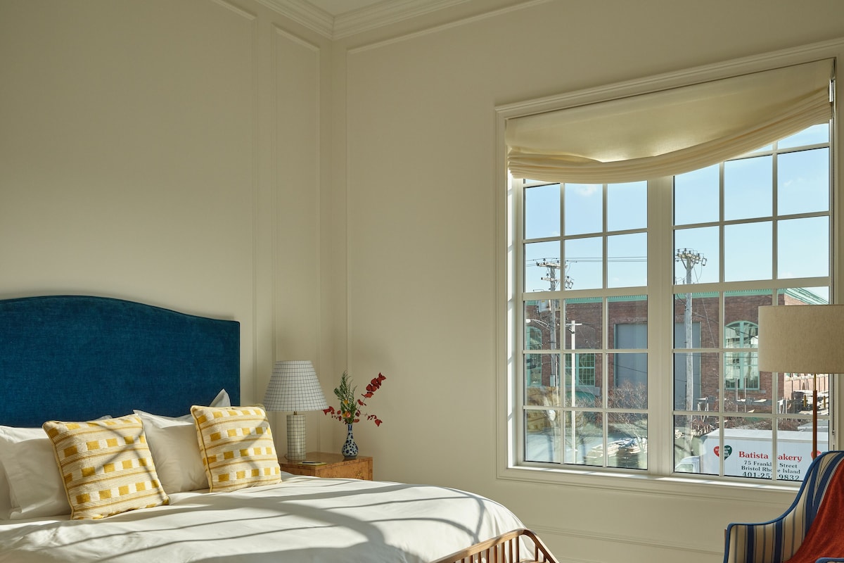 A bedroom with a large bed, a wooden bench, a chair, a nightstand with a lamp, and a window with a view of the outside.