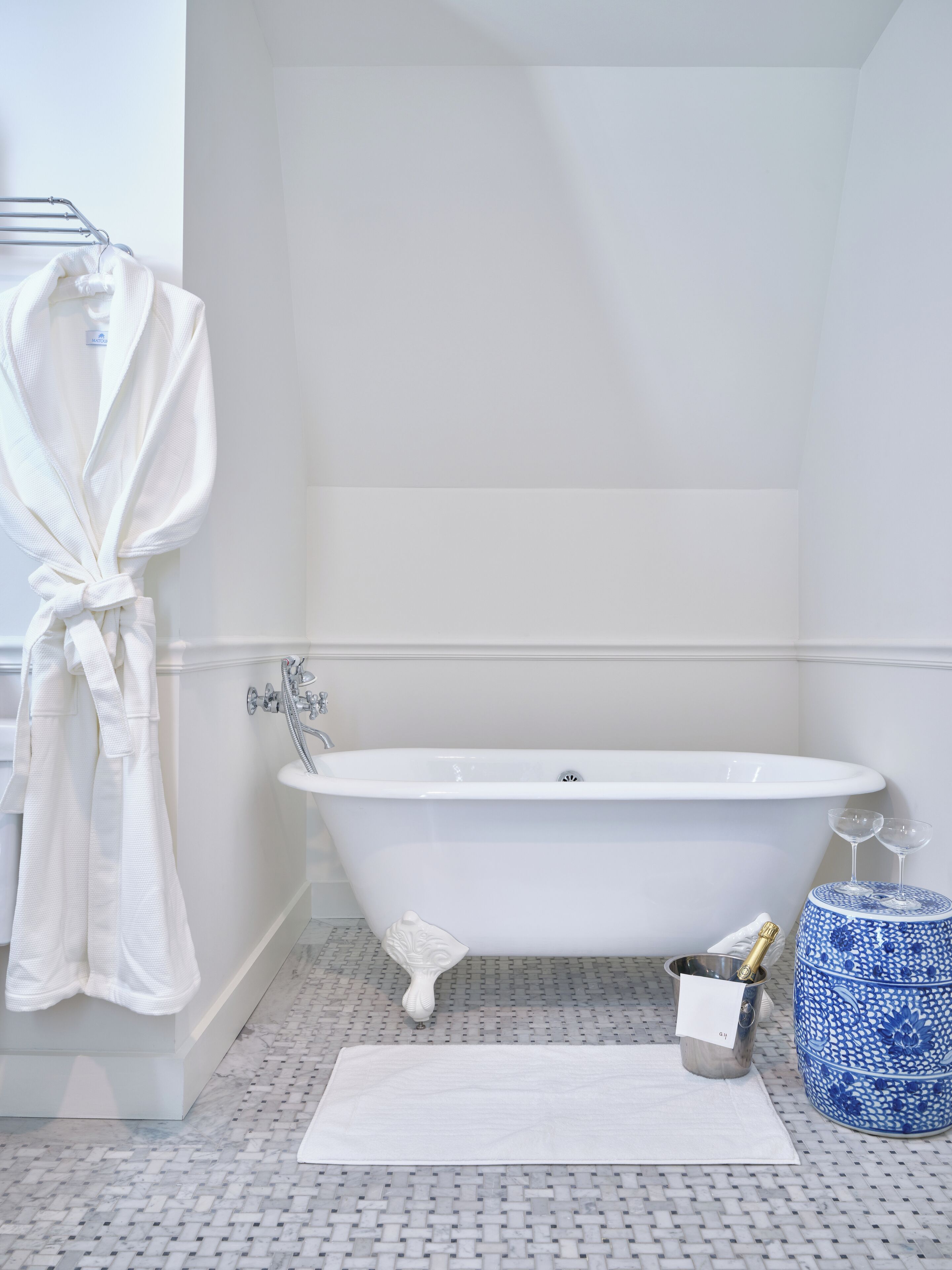 Une salle de bain avec une baignoire sur pieds, un peignoir blanc accroché à un porte-serviettes, un tapis de bain blanc, un petit objet décoratif et un vase bleu et blanc.