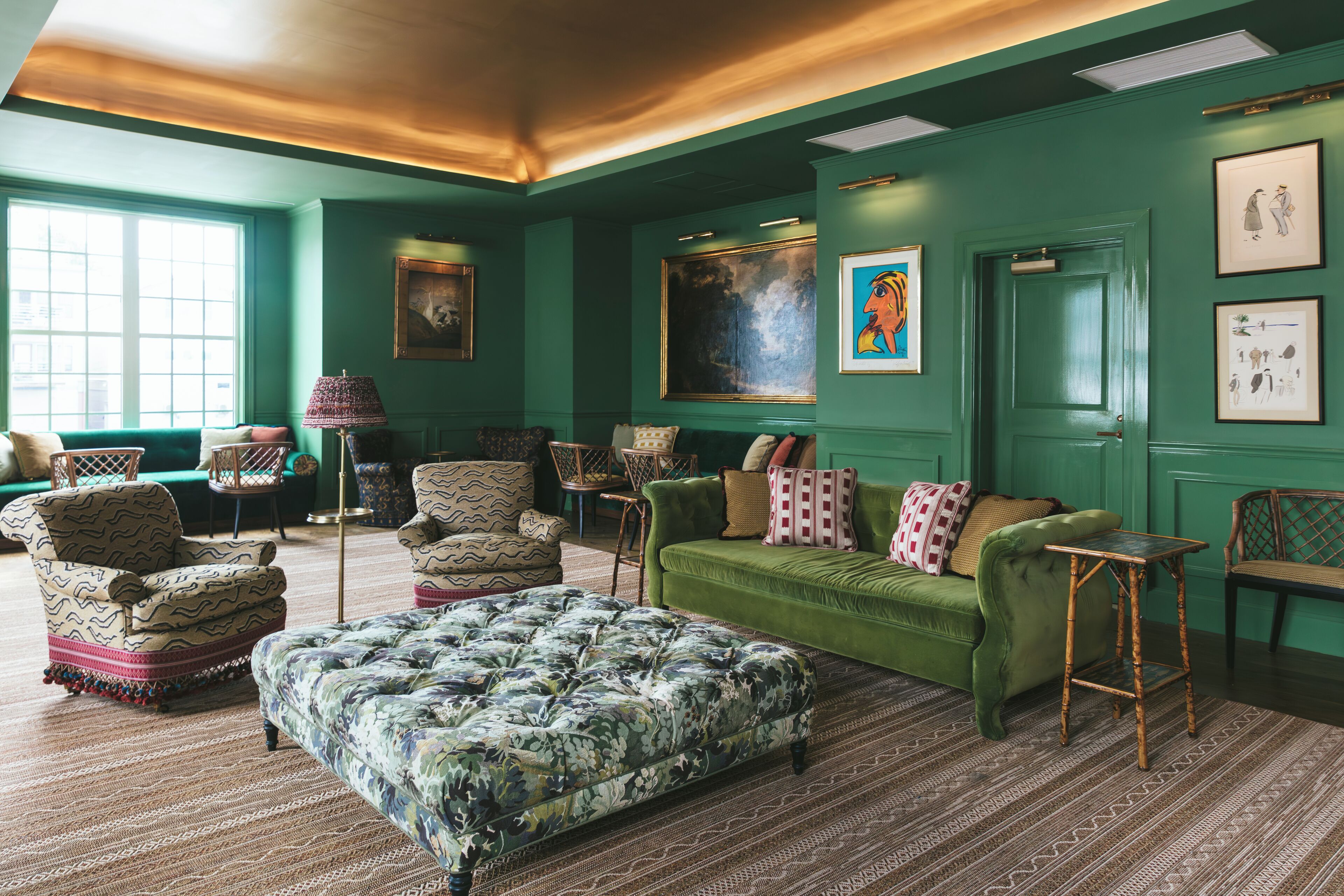 A room with a green wall, a green sofa, patterned armchairs, a tufted ottoman, and a small table.