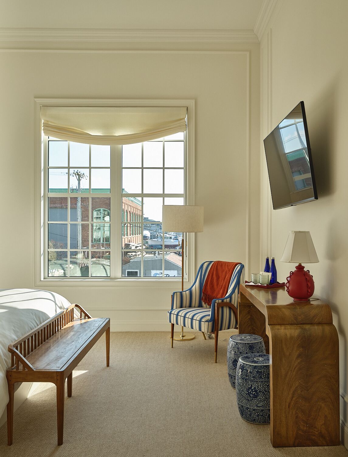 A hotel room with a bed, a desk with a lamp, a chair, a TV mounted on the wall, and a large window with a view of buildings.