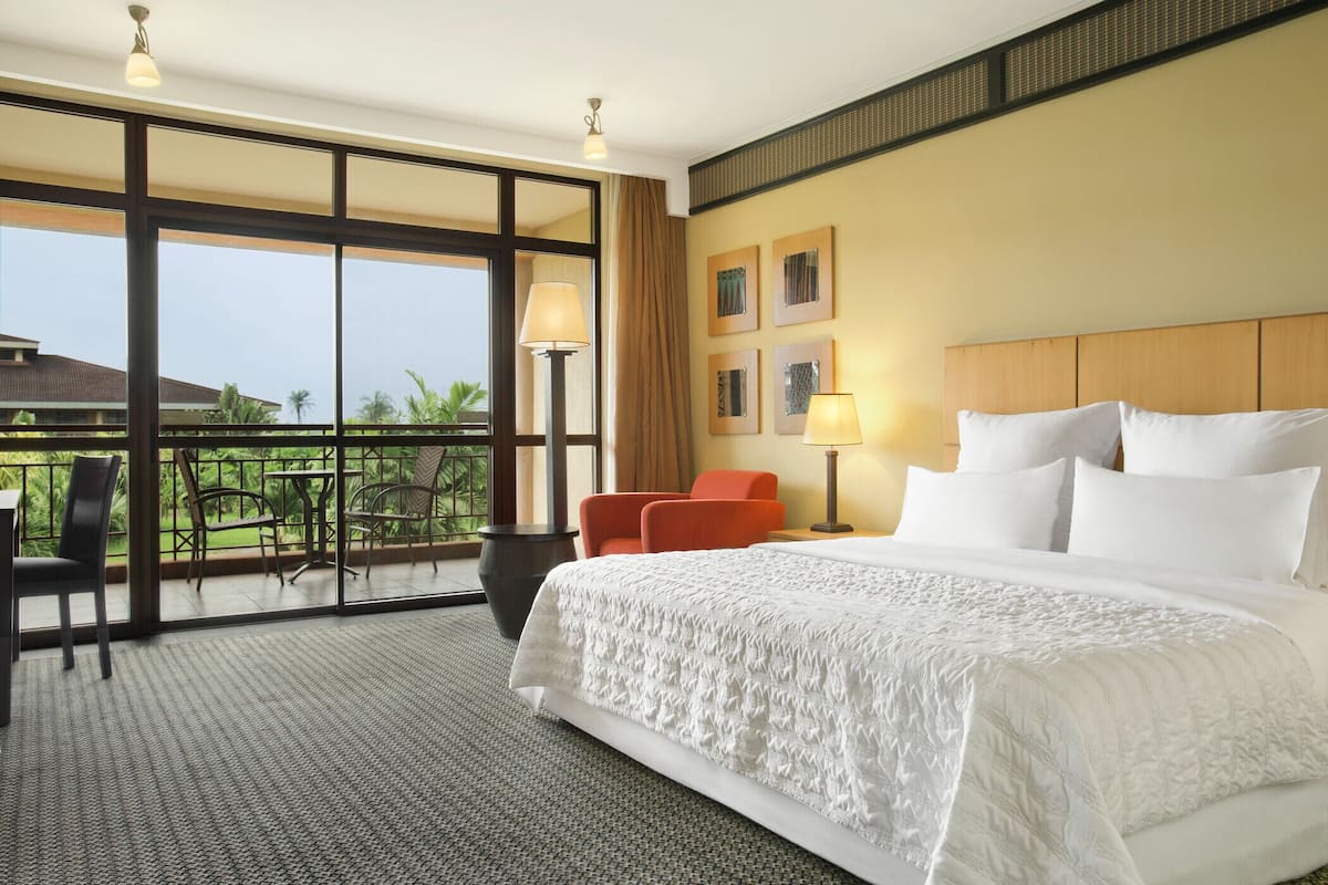 A hotel room with a large bed, a balcony with chairs, and a view of greenery.