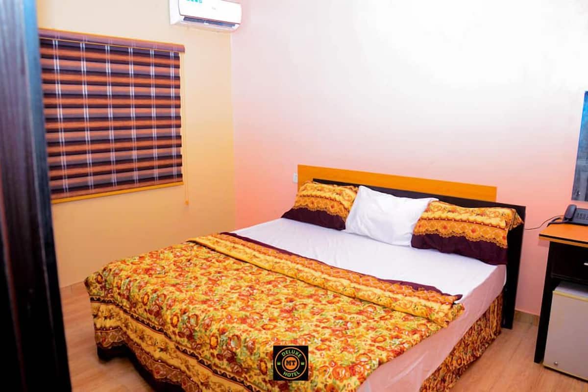 A hotel room with a bed, a bedside table, a lamp, a window with blinds, and a wall-mounted air conditioner.