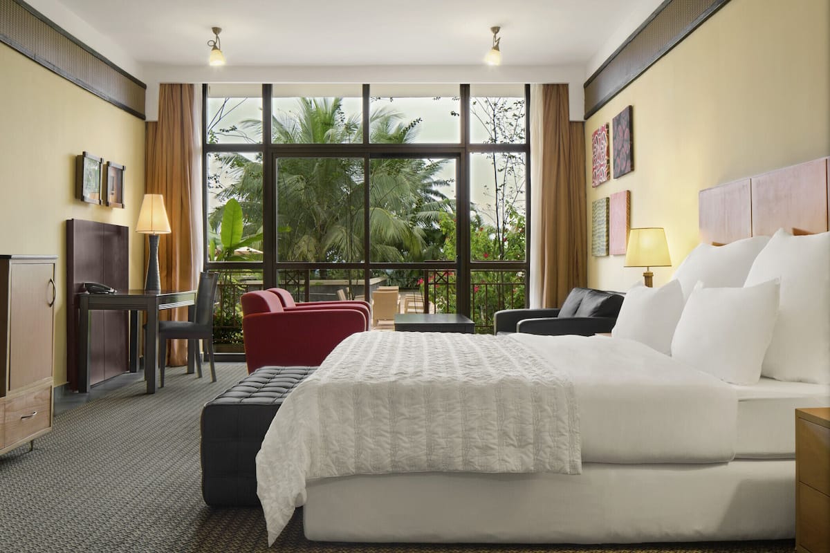 A hotel room with a large bed, a desk with a chair, a red chair, a small table, and a balcony with a view of palm trees.