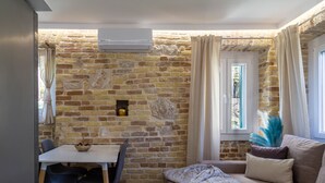 Ein gemütlicher Raum mit Steinwand, einem weißen Tisch mit Stühlen, einer Couch mit Kissen und einem Fenster mit Vorhängen.
