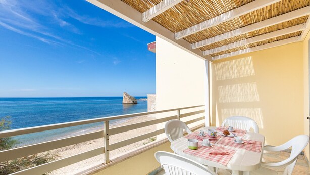 Un balcón con una mesa para dos, con vista a una playa de aguas azul turquesa y un promontorio rocoso.
