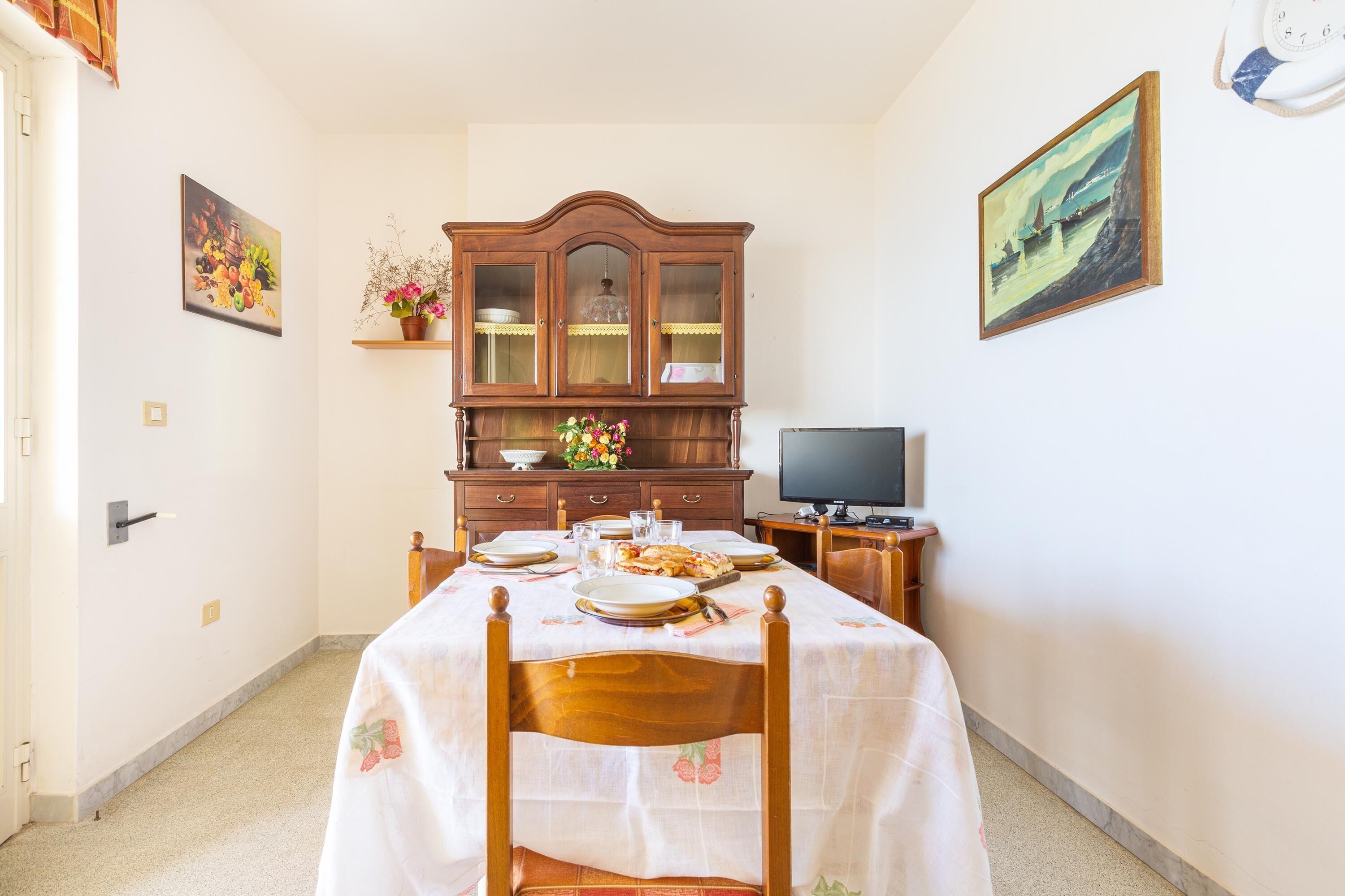 A dining area with a table set for two, a wooden cabinet with glass doors, a television on a stand, and a painting on the wall.