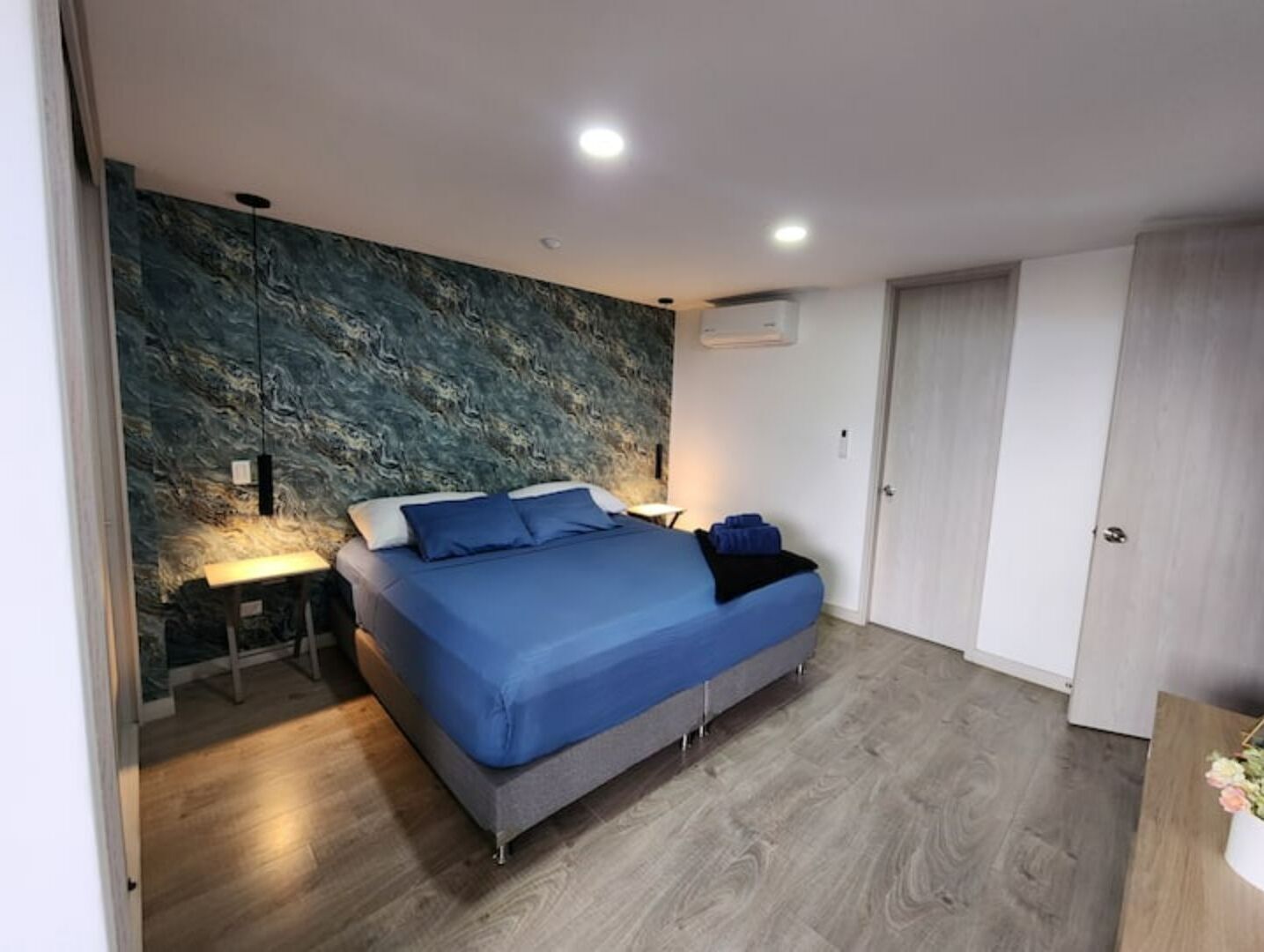 A modern bedroom with a large bed, a nightstand, a wall-mounted air conditioner, and a wooden floor.