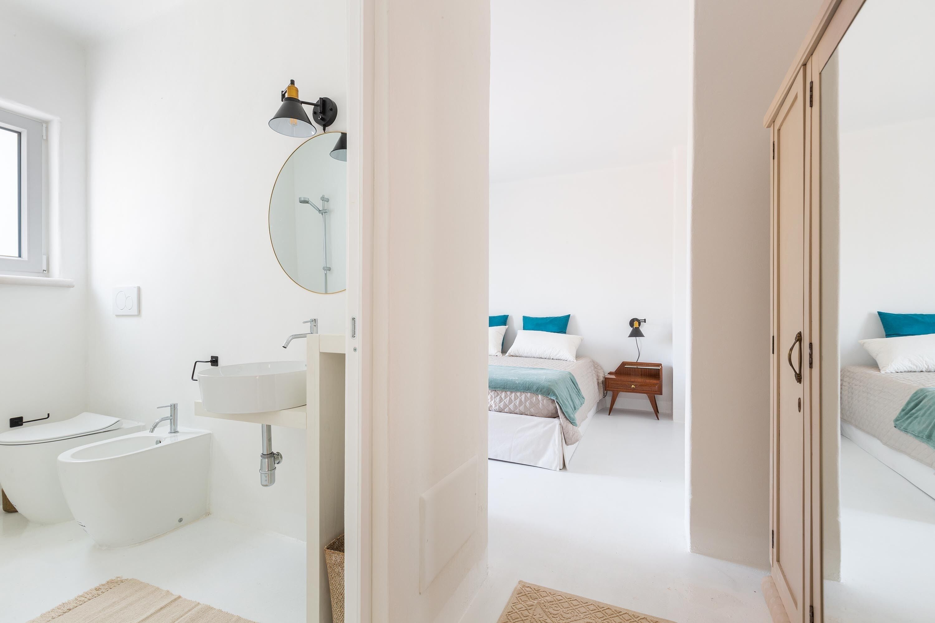 A modern bathroom with a bathtub, sink, and mirror. There is a bed with blue and white bedding in the background.