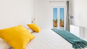 Ein Schlafzimmer mit einem Bett, zwei gelben Kissen, einer türkisblauen Decke, einem Fenster mit Blick auf Grünflächen und einem weißen Heizkörper.