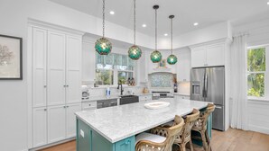A modern kitchen with white cabinets, a central island with bar stools, a stainless steel refrigerator, and decorative hanging lights. - Located in Captiva Village! Beautifully Designed Pool Home (Captiva)