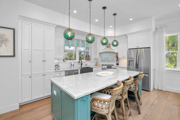 A modern kitchen with white cabinets, a central island with bar stools, a stainless steel refrigerator, and decorative hanging lights. - Located in Captiva Village! Beautifully Designed Pool Home (Captiva)