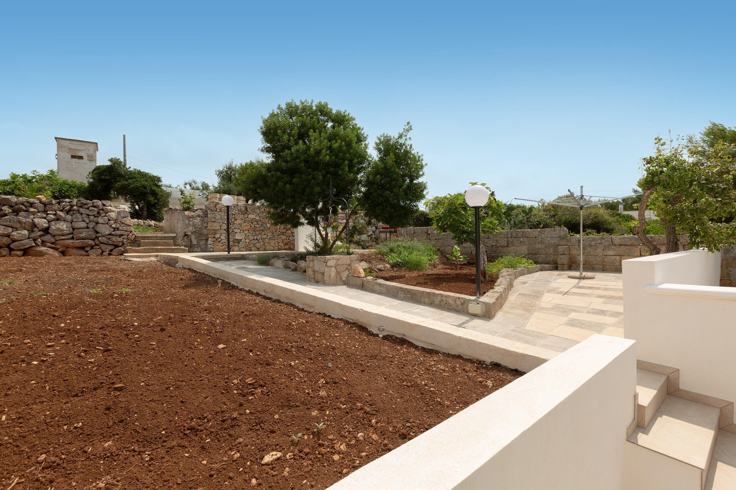 Un área exterior ajardinada con un muro de piedra, un sendero pavimentado y un edificio blanco con escalones.